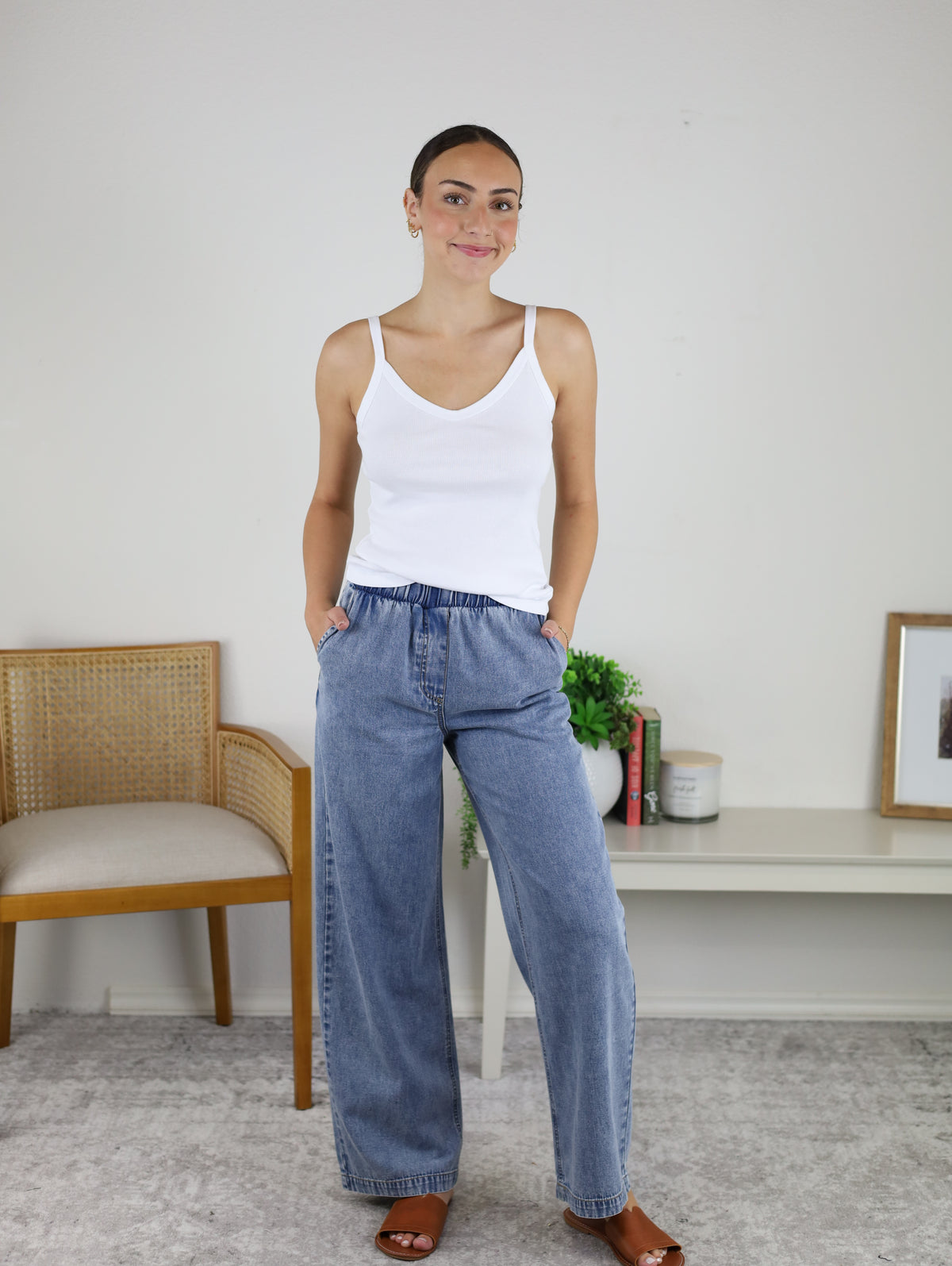 A woman stands indoors, smiling with her hands in her pockets, wearing a white tank top and the Marigold Acid Wash Wide Leg Denim. She pairs the look with brown sandals. In the background are a wooden chair, a shelf with books, a plant, and a framed picture.