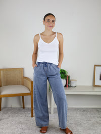 A woman stands indoors, smiling with her hands in her pockets, wearing a white tank top and the Marigold Acid Wash Wide Leg Denim. She pairs the look with brown sandals. In the background are a wooden chair, a shelf with books, a plant, and a framed picture.