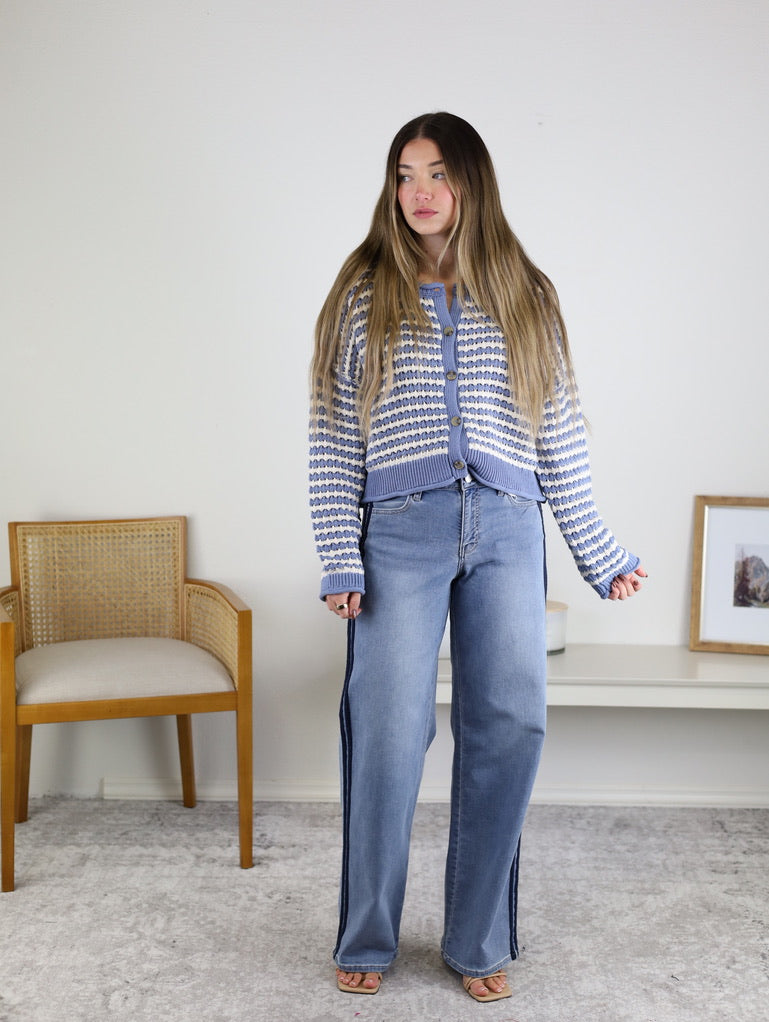 A woman with long hair wears the Bellamy Textured Striped Crochet Cardigan and wide-leg jeans, standing indoors beside a wooden chair and a white shelf with framed photos.
