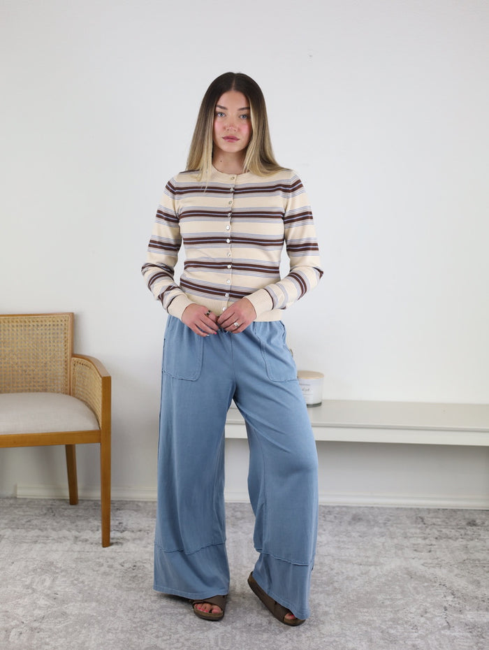 A woman stands indoors wearing the Sausalito Soft Striped Cardigan and loose blue pants. She faces forward with her arms by her sides, while a wooden chair and white shelves are visible in the background.