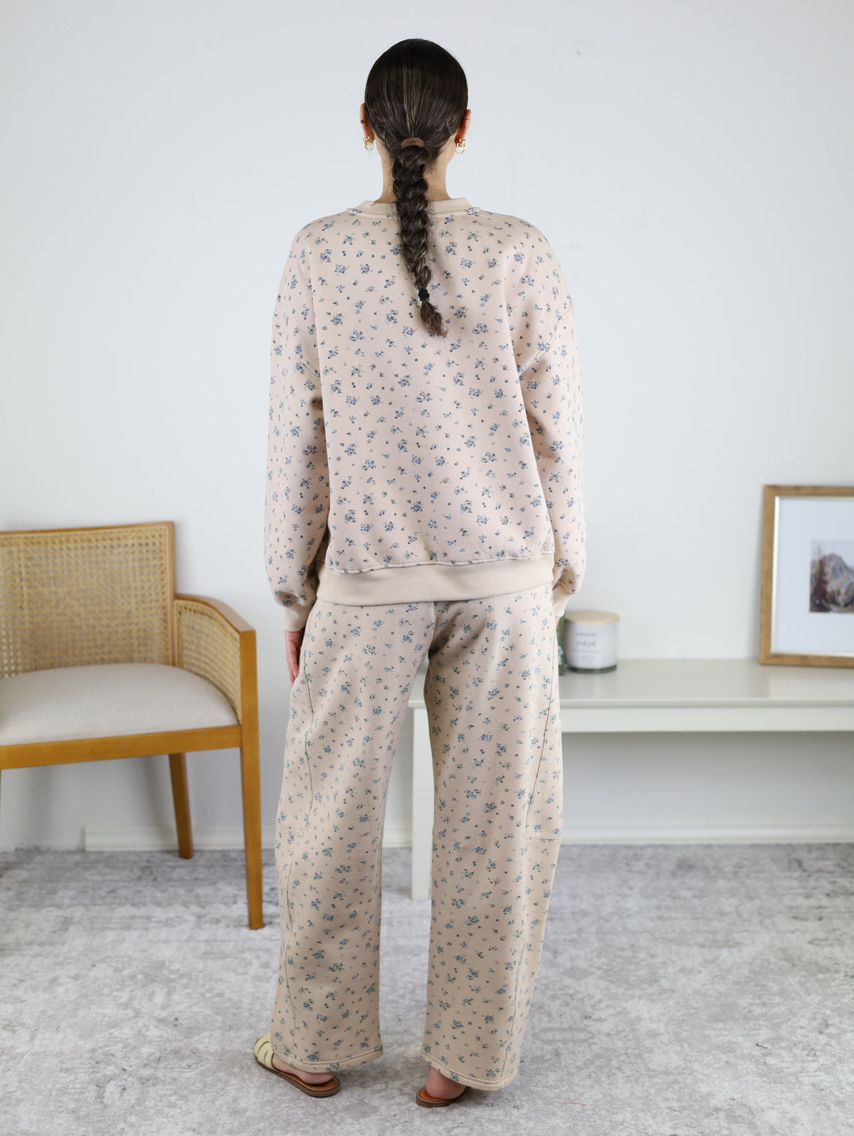 A person with a long braid, wearing the Journey Floral Boxy Crewneck Lounge Set in khaki, stands facing away in a minimalist room featuring a chair, shelf, candle, and framed photo.