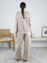 A person with a long braid, wearing the Journey Floral Boxy Crewneck Lounge Set in khaki, stands facing away in a minimalist room featuring a chair, shelf, candle, and framed photo.