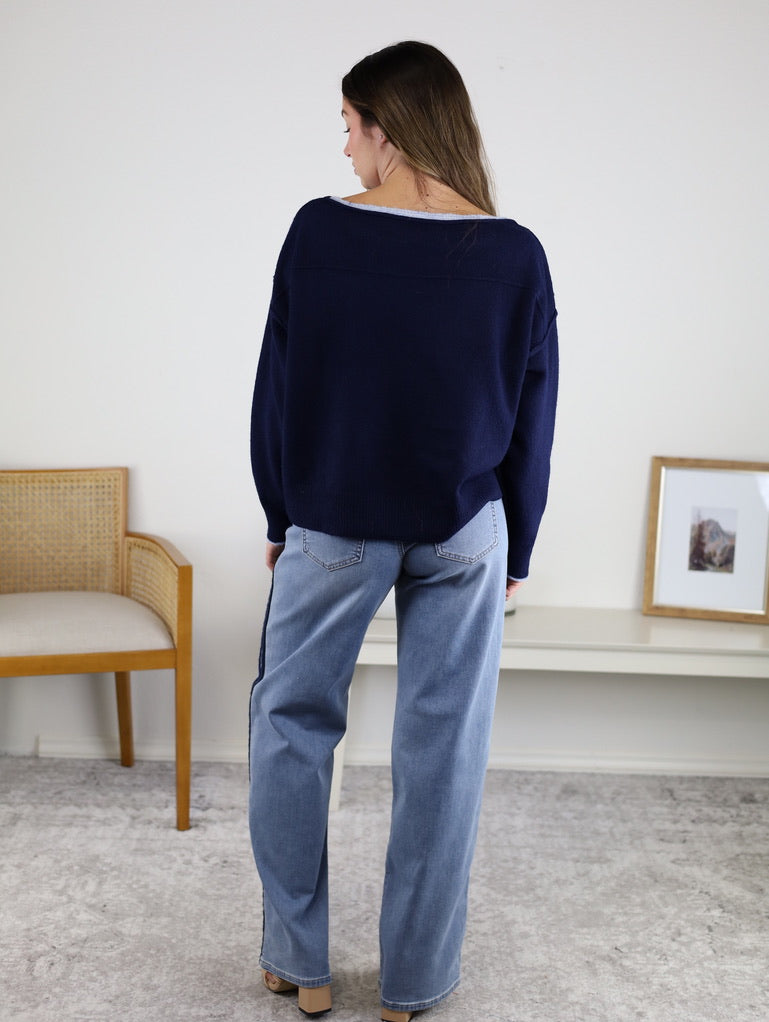 A woman stands indoors facing away, wearing the Bell Contrast Boat Neck Sweater in navy and light blue jeans. A wooden chair and a framed picture on a white bench are in the background.