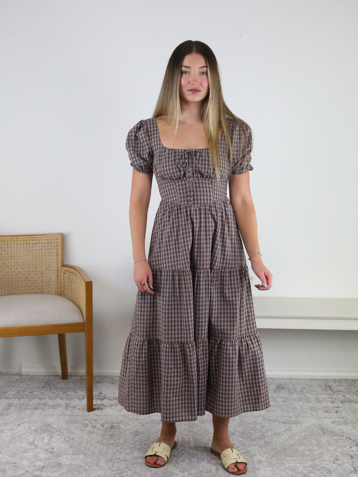 A woman stands indoors wearing the Fable Puff Sleeve Midi Dress—a brown plaid midi dress featuring puff sleeves and a tiered skirt—paired with cream sandals and subtle jewelry. To her left is a wooden chair with a cushioned seat, and to her right is a white bench.