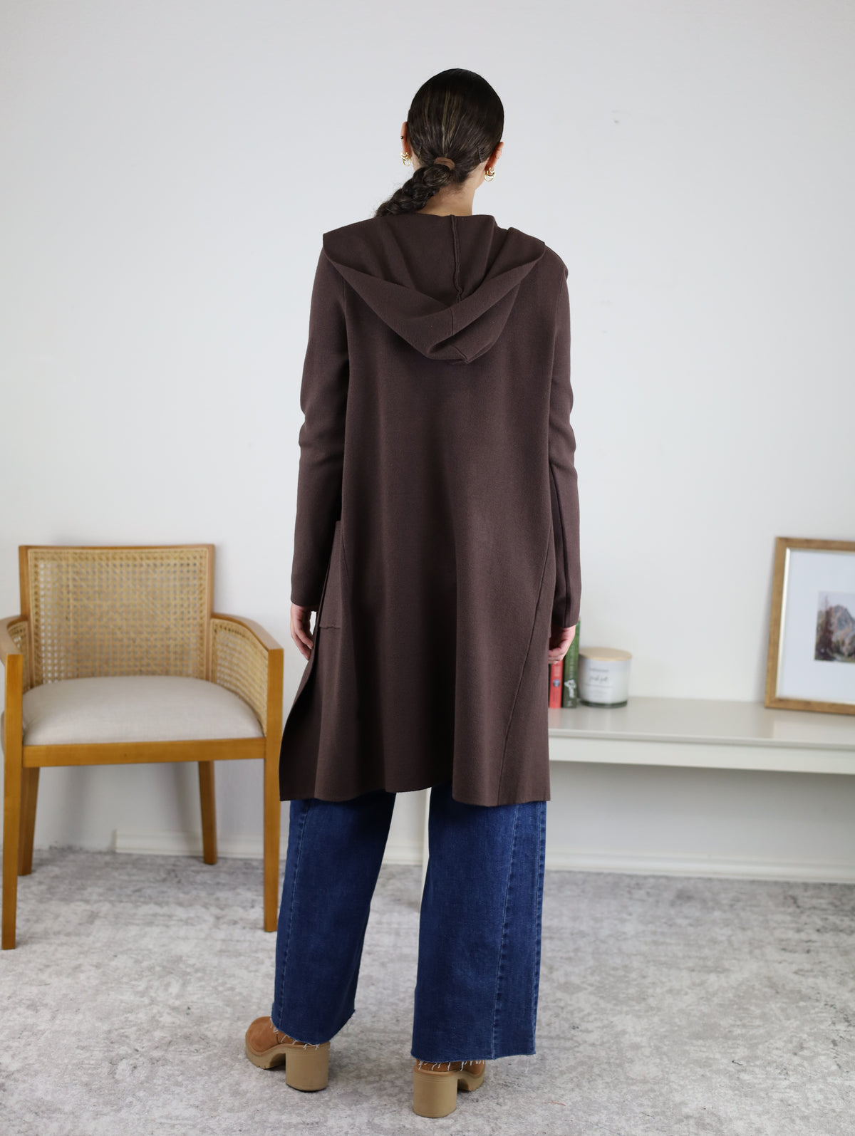 A person stands facing away, wearing the Pinch Me Long Cardigan in a warm brown hue, paired with wide-leg blue jeans and tan shoes. The room features a chair, a low table with candles, and a framed photo on a white wall—creating an atmosphere perfect for cozy layering.