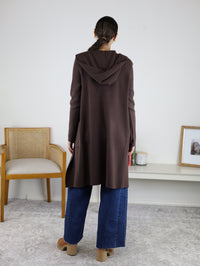 A person stands facing away, wearing the Pinch Me Long Cardigan in a warm brown hue, paired with wide-leg blue jeans and tan shoes. The room features a chair, a low table with candles, and a framed photo on a white wall—creating an atmosphere perfect for cozy layering.