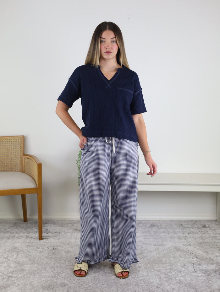A woman stands indoors wearing a navy short-sleeve top and the Willa Gingham Ruffle Hem Cropped Pants. She has straight hair and is barefoot, posing with one hand on her hip. A wooden chair and white bench are in the background, highlighting her stylish choice of women’s spring pants.