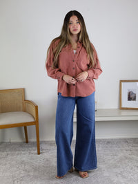 Woman with long hair stands indoors wearing the Larkin Rib Knit Button Up in pink, wide-leg blue jeans, and sandals. A wooden chair and framed picture are on the light grey carpet background.