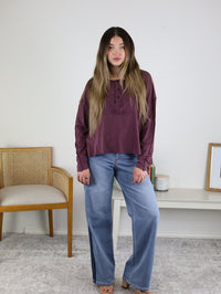A woman with long brown hair wears the Ester Raw Seam Henley in maroon and wide-leg blue jeans. She stands indoors by a beige chair and a white shelf, against a plain white background.