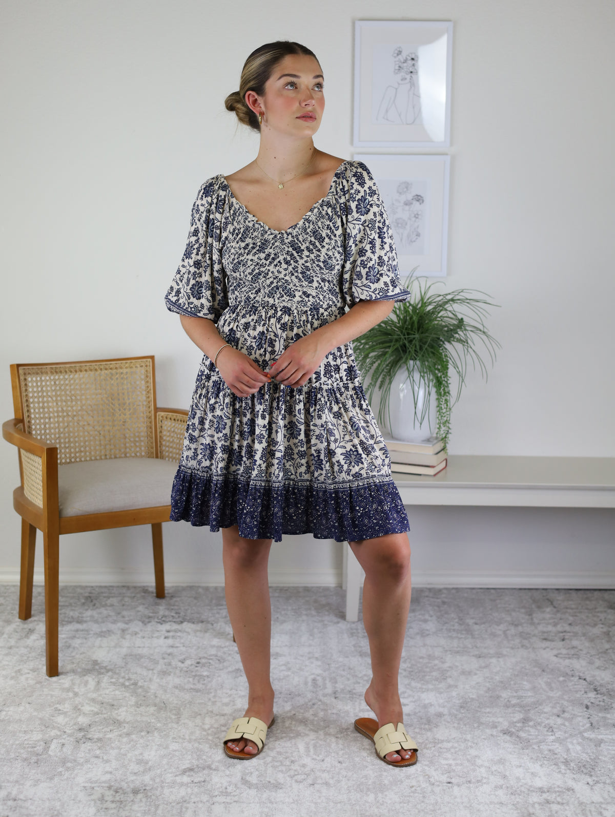 A woman wearing a navy and cream floral-print Leslie Smocked Mini Dress with a V-neckline, elbow-length puff sleeves, and a lace-up back, paired with cream slide sandals.