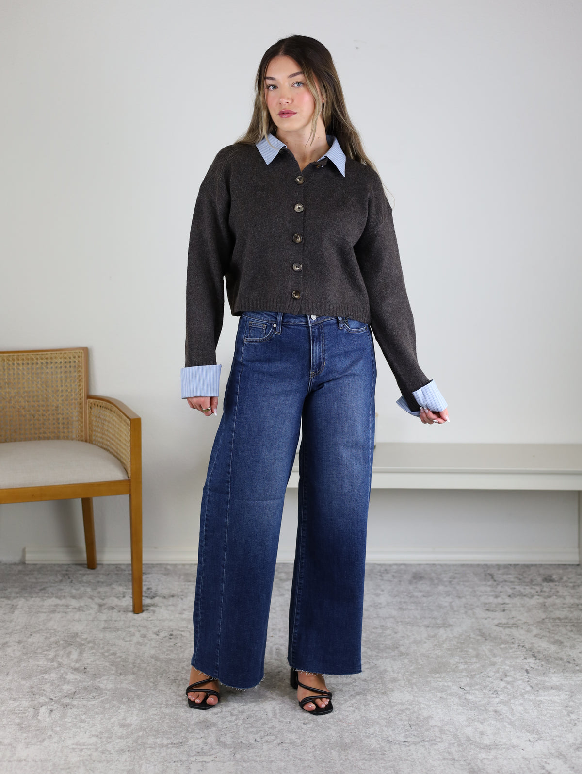 A woman stands indoors wearing the Mani Stripe Trim Button Down Cardigan over a blue collared shirt, wide-leg jeans, and black heels. A wooden chair with a cushion sits to her left, and a white shelf is visible behind her.