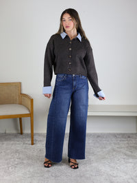 A woman stands indoors wearing the Mani Stripe Trim Button Down Cardigan over a blue collared shirt, wide-leg jeans, and black heels. A wooden chair with a cushion sits to her left, and a white shelf is visible behind her.