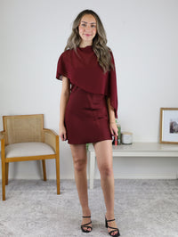 A woman models the Amy Mini Dress with Sheer Cape, a short, maroon satin party dress with a mock neck and a sheer cape overlay.