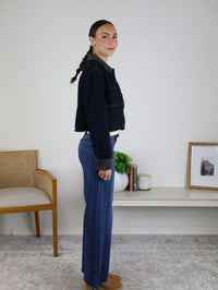 A woman with a braid stands sideways indoors, wearing the Autumn Leopard Collar Denim Shacket and blue jeans. She is beside a wooden chair and a white shelf adorned with books, a plant, and a framed picture.