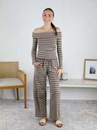 A woman stands indoors, smiling, wearing the Linn Off Shoulder Top Barrel Pant Set in brown and white stripes, paired with beige sandals. She is in a bright, minimally decorated room, radiating effortless loungewear style.
