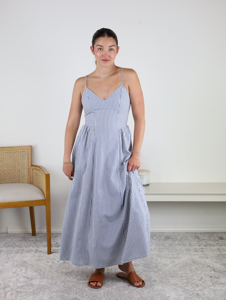 Wearing the Molly Striped Maxi Dress with adjustable straps, this woman stands indoors on a light carpet with a neutral background, wooden chair, and bench beside her. She pairs the blue and white striped dress with brown sandals and has her hair tied back.