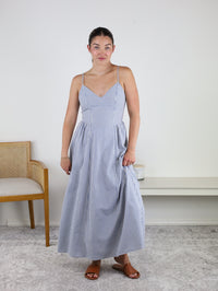 Wearing the Molly Striped Maxi Dress with adjustable straps, this woman stands indoors on a light carpet with a neutral background, wooden chair, and bench beside her. She pairs the blue and white striped dress with brown sandals and has her hair tied back.