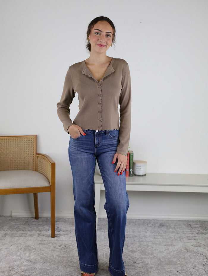 A woman stands indoors, smiling with one hand in her pocket, wearing the Juniper Scalloped Cardigan in taupe and blue jeans. Behind her are a wooden chair and a white table adorned with books and a small plant.