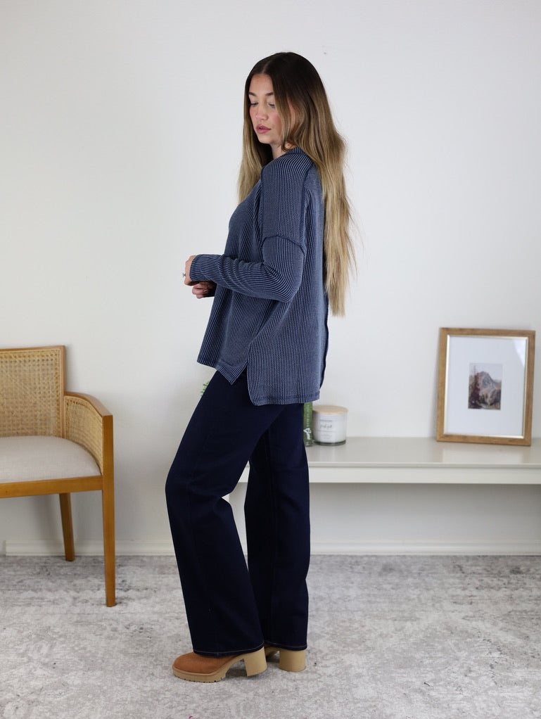 A woman with long hair stands sideways in a room wearing a blue Finley Sweater, dark pants, and tan shoes. Behind her are a chair, shelf with a candle, and framed picture on a white wall.