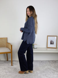 A woman with long hair stands sideways in a room wearing a blue Finley Sweater, dark pants, and tan shoes. Behind her are a chair, shelf with a candle, and framed picture on a white wall.
