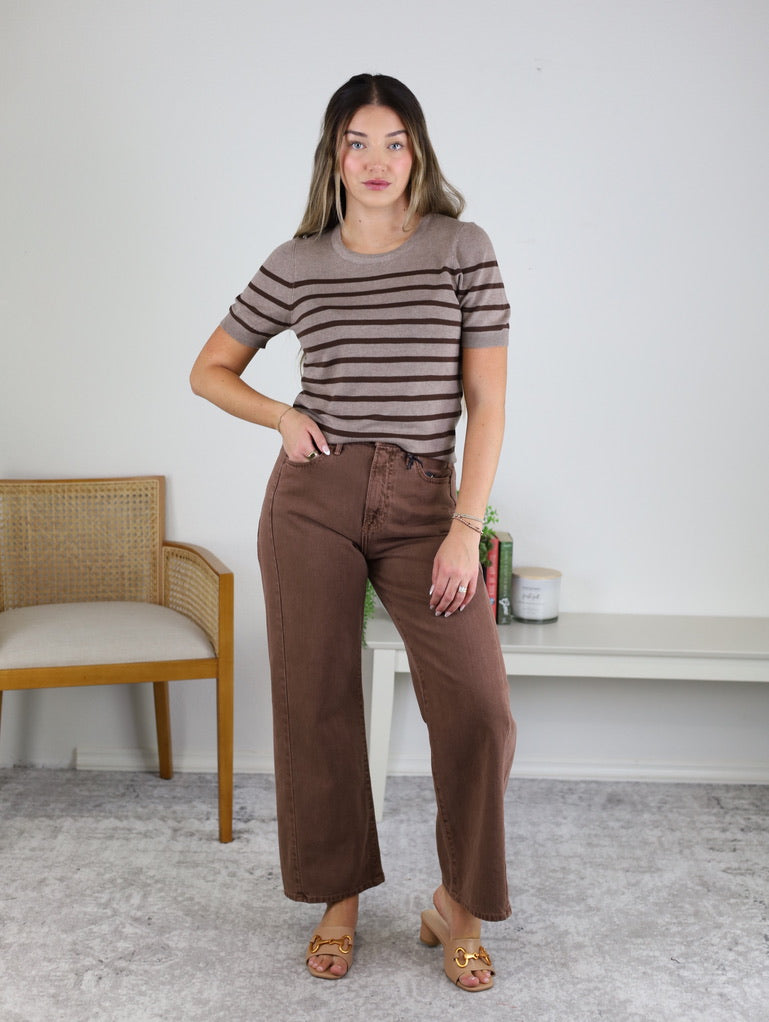A woman stands indoors wearing the Alex Striped Shortsleeve Sweater Top, paired with wide-leg brown pants and tan sandals. She poses with one hand on her hip and the other relaxed. In the background, there’s a chair and a table adorned with decor.