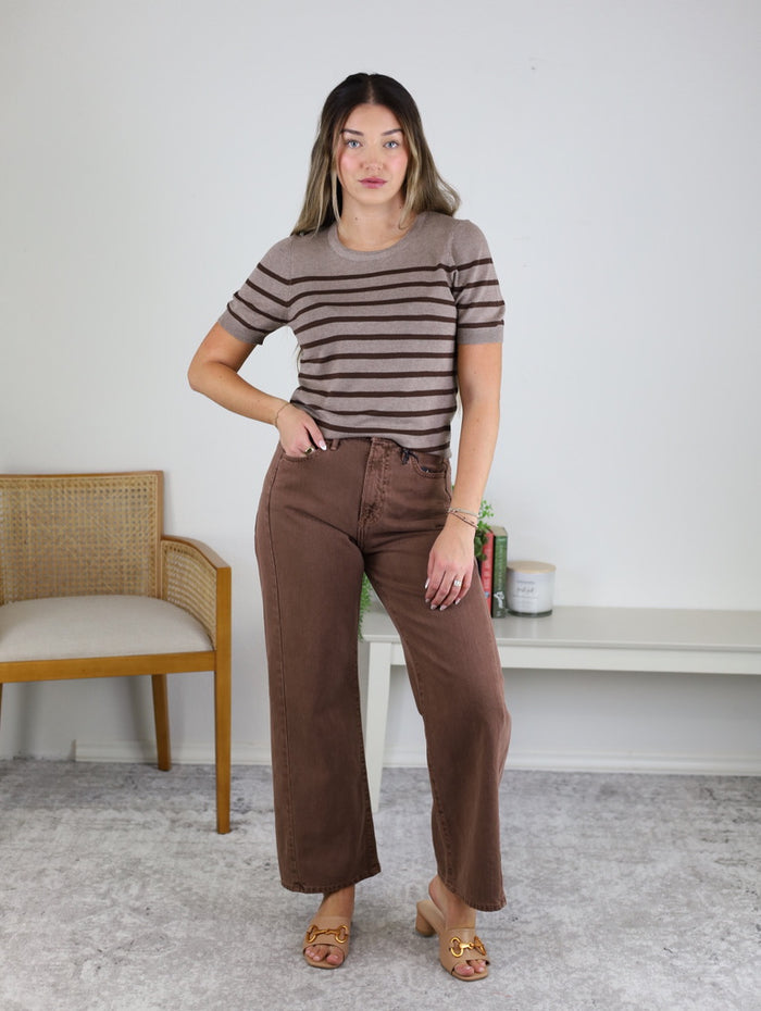 A woman stands indoors wearing the Raj High Rise Twist Seam Barrel Jean paired with a vintage brown denim striped top and tan heeled sandals. She is positioned in front of a white wall, beside a wooden chair and a white table adorned with candles and decor.