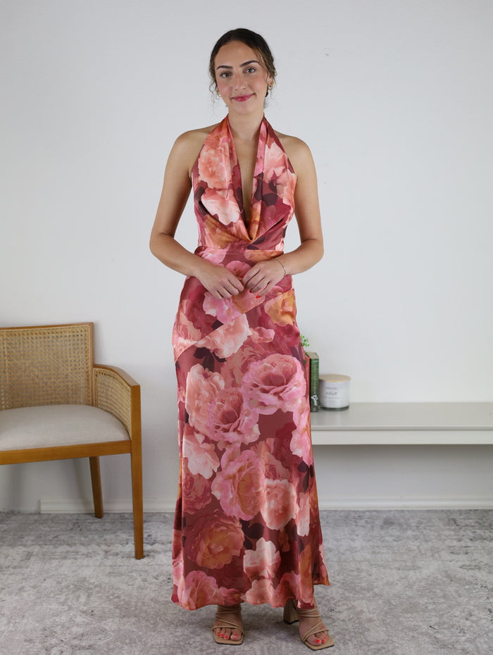 A woman wearing the Presley Floral Draped Halter Neck Maxi Dress stands indoors on a gray carpet, beside a wooden chair and a white bench adorned with a candle and plant.