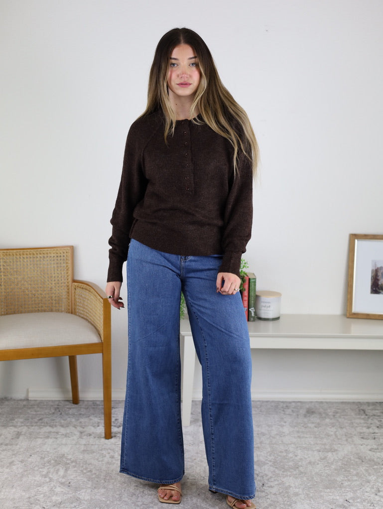 A woman with long hair wears the Theo Sweater and wide-leg blue jeans, standing indoors on a gray carpet near a wooden chair and a white shelf with books, a candle, and framed art.