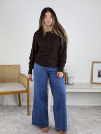 A woman with long hair wears the Theo Sweater and wide-leg blue jeans, standing indoors on a gray carpet near a wooden chair and a white shelf with books, a candle, and framed art.