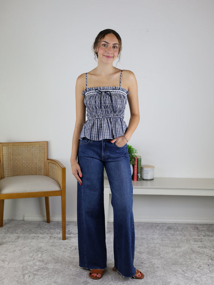 A woman stands indoors, smiling with one hand in her pocket, wearing the Lonnie Smocked Gingham Peplum Top with adjustable spaghetti straps and wide-leg jeans. In the background, there is a chair, a small table, and a plant.
