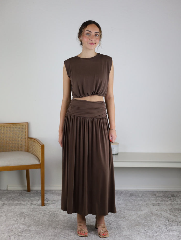 A woman stands indoors wearing the Harlow Bubble Top w/ Skirt, a sleeveless brown bubble hem crop top paired with the matching skirt and sandals. She smiles slightly, with a wooden chair and white bench positioned in front of a plain white wall in the background.