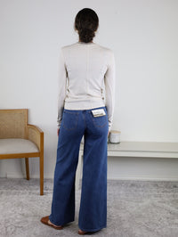 A person with long hair in the Josette Ribbed Classic Crewneck and wide-leg blue jeans stands facing a wall. A woven chair is on the left, and a white bench with a candle and decor is on the right.