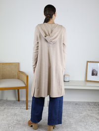 A woman stands indoors with her back to the camera, showcasing cozy layering in the Pinch Me Long Cardigan—a beige hooded style—paired with wide-leg blue jeans and brown shoes. A chair and a low table with decor appear in the background.