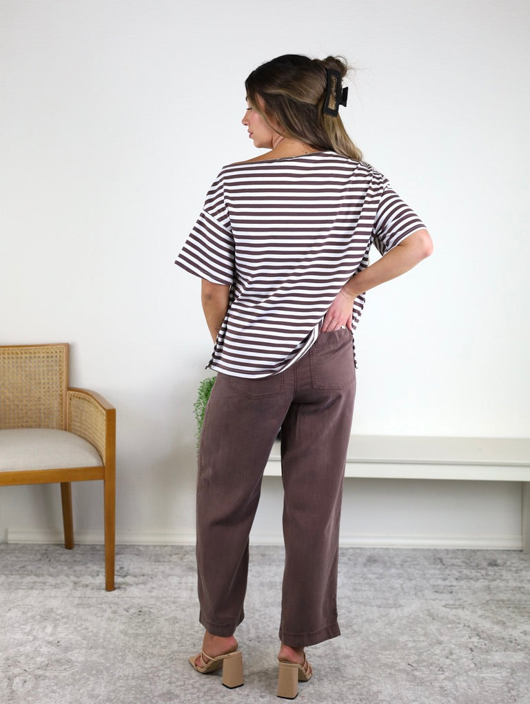 A woman stands indoors facing away, wearing the Hayden Asymmetrical Off-Shoulder Boxy Cotton Tee in black-and-white stripes, paired with brown pants and heeled sandals. She has a hair clip in her hair and is positioned next to a wooden chair and a white bench against a white wall.