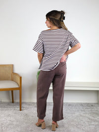 A woman stands indoors facing away, wearing the Hayden Asymmetrical Off-Shoulder Boxy Cotton Tee in black-and-white stripes, paired with brown pants and heeled sandals. She has a hair clip in her hair and is positioned next to a wooden chair and a white bench against a white wall.