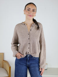 A woman with long, dark braided hair stands indoors smiling with one hand in her pocket, wearing the Beth Lightweight Cardigan in beige and blue jeans. A wooden chair and a white table are in the background.