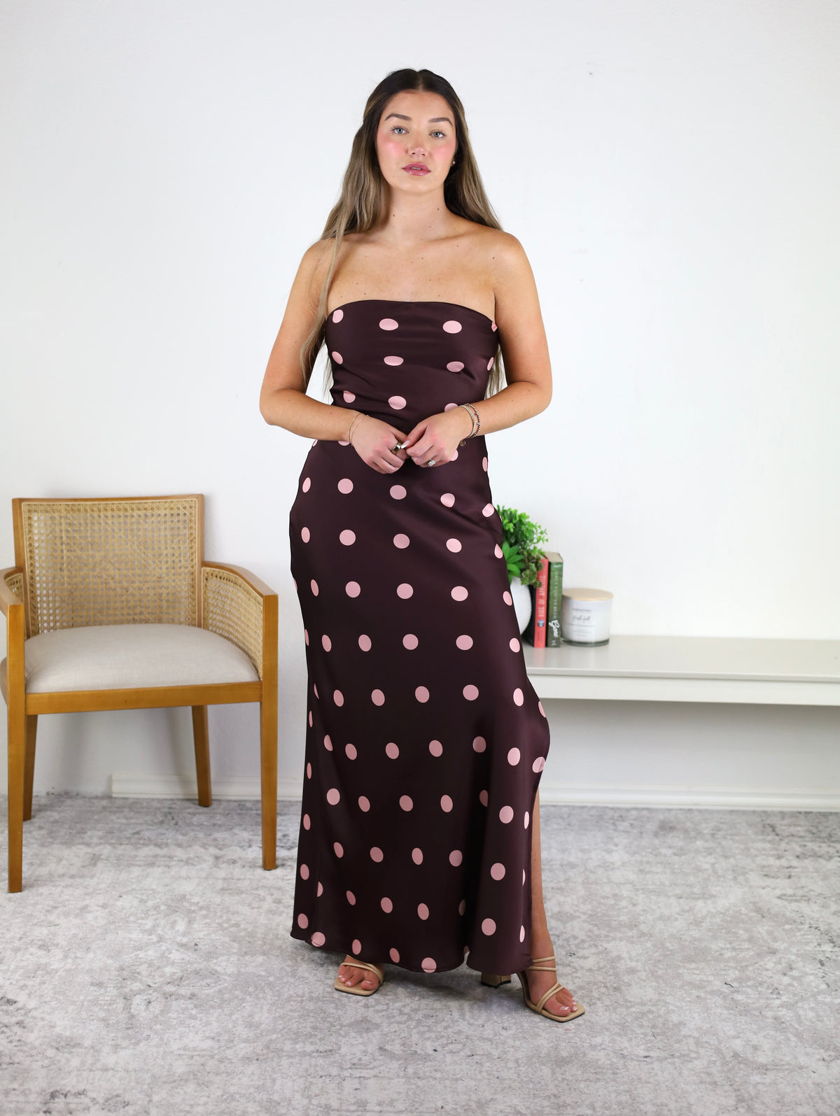 A woman stands indoors wearing the Polka Dot Satin Strapless Maxi Dress, which features a brown satin fabric adorned with large pink polka dots. She has long hair, a neutral expression, and is holding her hands together. In the background, there is a chair, table, books, and a plant.