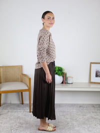 A woman stands sideways indoors, smiling as she models the Maci Satin Crinkle Pleated Maxi Skirt in a deep, dark hue paired with a beige polka-dot sweater—an ideal ensemble for a cozy fall wardrobe. In the background, there’s a chair, plant, books, and framed photo.