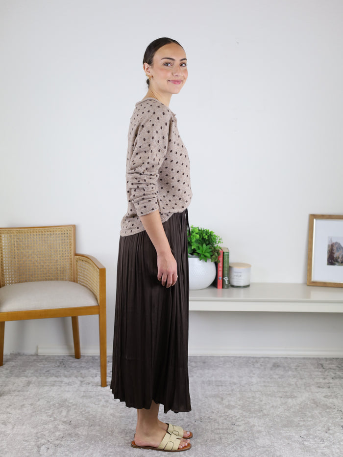 A woman stands sideways indoors, smiling as she models the Maci Satin Crinkle Pleated Maxi Skirt in a deep, dark hue paired with a beige polka-dot sweater—an ideal ensemble for a cozy fall wardrobe. In the background, there’s a chair, plant, books, and framed photo.