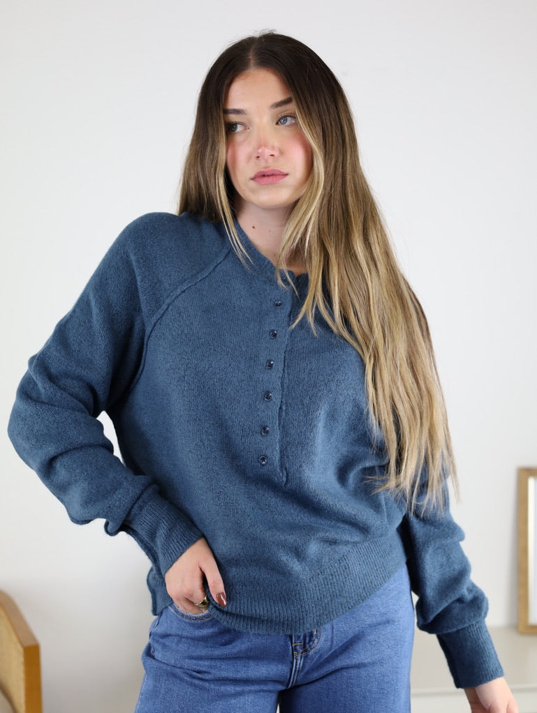 A woman with long, light brown hair wears a relaxed fit Theo Sweater in blue with jeans, standing indoors against a light background and looking slightly to the side.