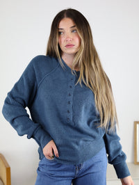 A woman with long, light brown hair wears a relaxed fit Theo Sweater in blue with jeans, standing indoors against a light background and looking slightly to the side.