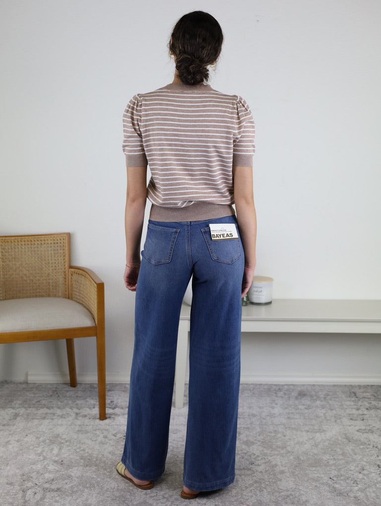 A woman stands indoors facing away, wearing the Ace Puff Striped Sweater and wide-leg blue jeans. There is a wooden chair on the left and a white table with a candle and container on the right.