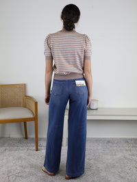 A woman stands indoors facing away, wearing the Ace Puff Striped Sweater and wide-leg blue jeans. There is a wooden chair on the left and a white table with a candle and container on the right.