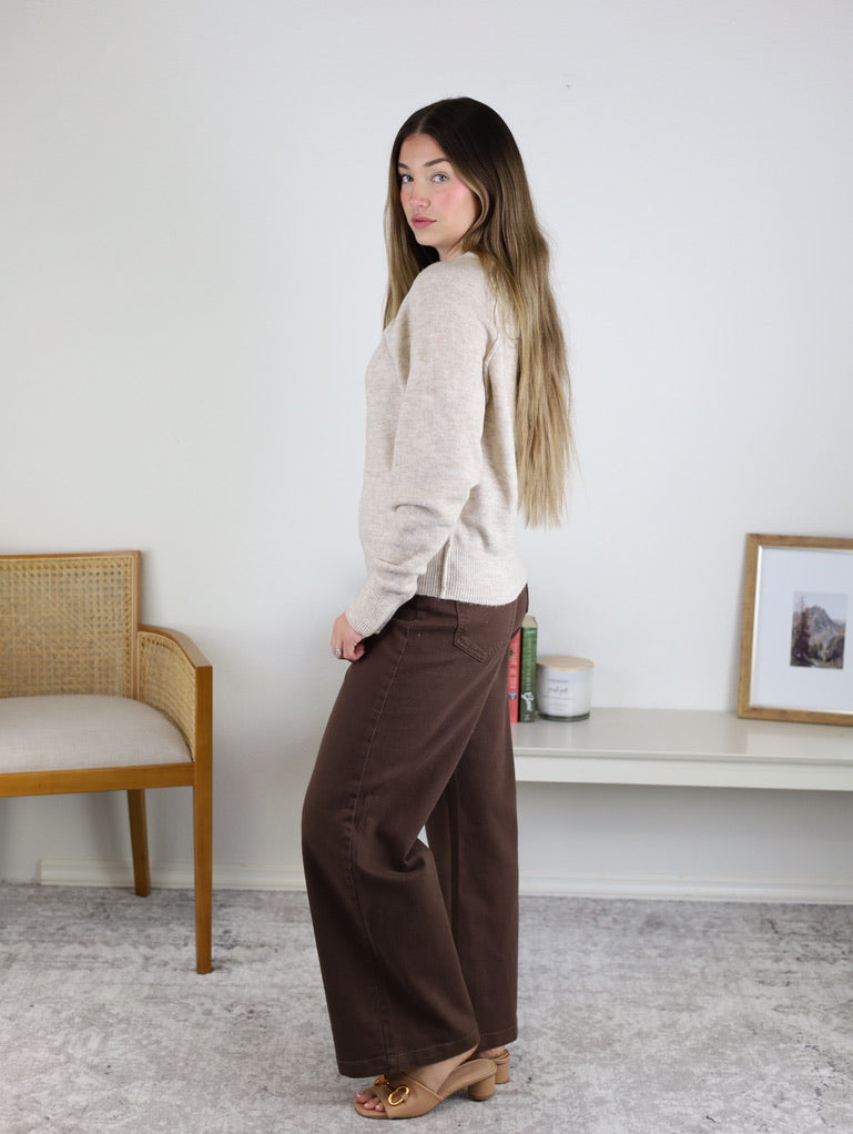 A woman with long brown hair stands sideways indoors, wearing a Theo Sweater with brown wide-leg pants and tan heels, near a wooden chair and a shelf with books and a framed photo.
