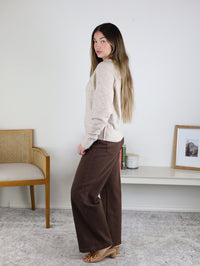 A woman with long brown hair stands sideways indoors, wearing a Theo Sweater with brown wide-leg pants and tan heels, near a wooden chair and a shelf with books and a framed photo.