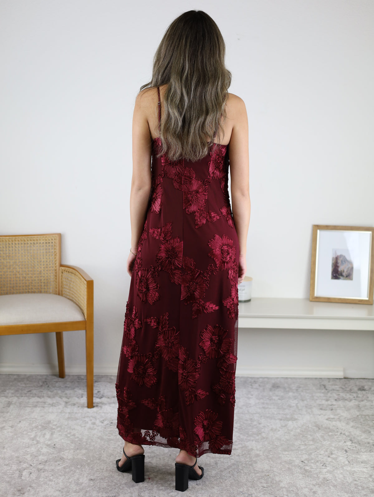 A person stands facing away in a dark red, floor-length Axel Applique Mesh Maxi Dress with romantic floral appliqué detailing and black heeled sandals.