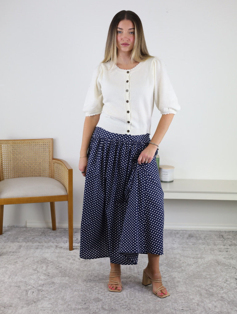 A woman stands indoors wearing the Monica Short Sleeve Cardigan, a white short sleeve cardigan with metallic buttons, paired with a long navy blue skirt featuring white polka dots. She has light brown hair, a neutral expression, and is wearing beige sandals. A chair and table are visible in the background.