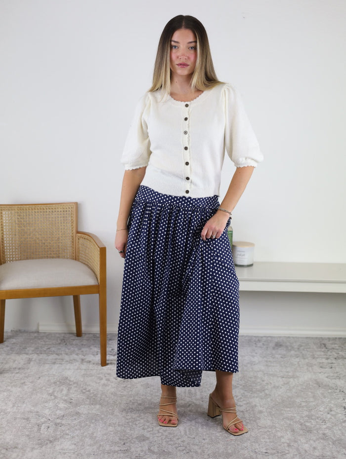 A woman stands indoors wearing the Monica Short Sleeve Cardigan, a white short sleeve cardigan with metallic buttons, paired with a long navy blue skirt featuring white polka dots. She has light brown hair, a neutral expression, and is wearing beige sandals. A chair and table are visible in the background.