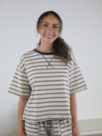A person with curly hair smiles, wearing the Lula Striped Pant Set—a cream and navy ensemble featuring a matching shirt and wide-leg pants—while standing against a plain white background.