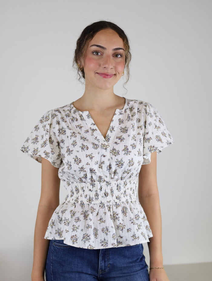 A woman smiles at the camera against a plain background, wearing the Tovar Floral Blouse—a white, short-sleeved peplum top with a cinched waist—paired with blue jeans.
